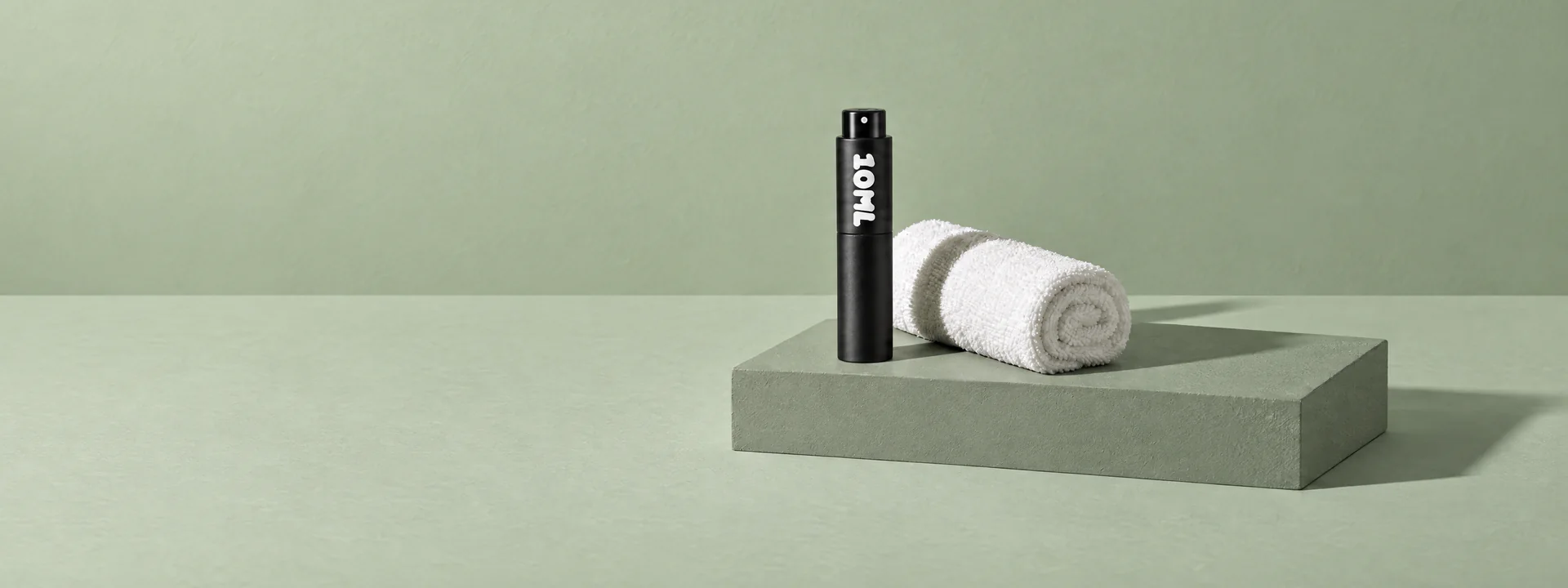 A black 10ML atomiser beside a rolled white towel on a sage-green plinth